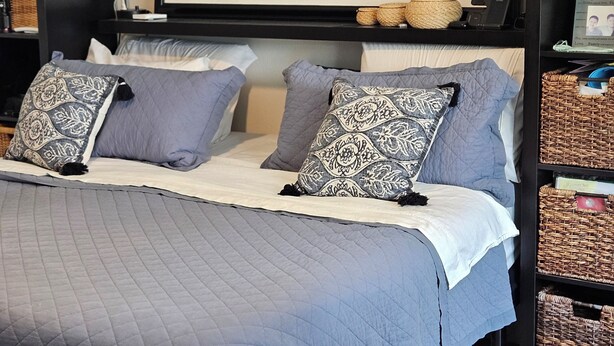 Una cama bien hecha con una colcha acolchada, dos almohadas estampadas y un cuadro enmarcado encima.