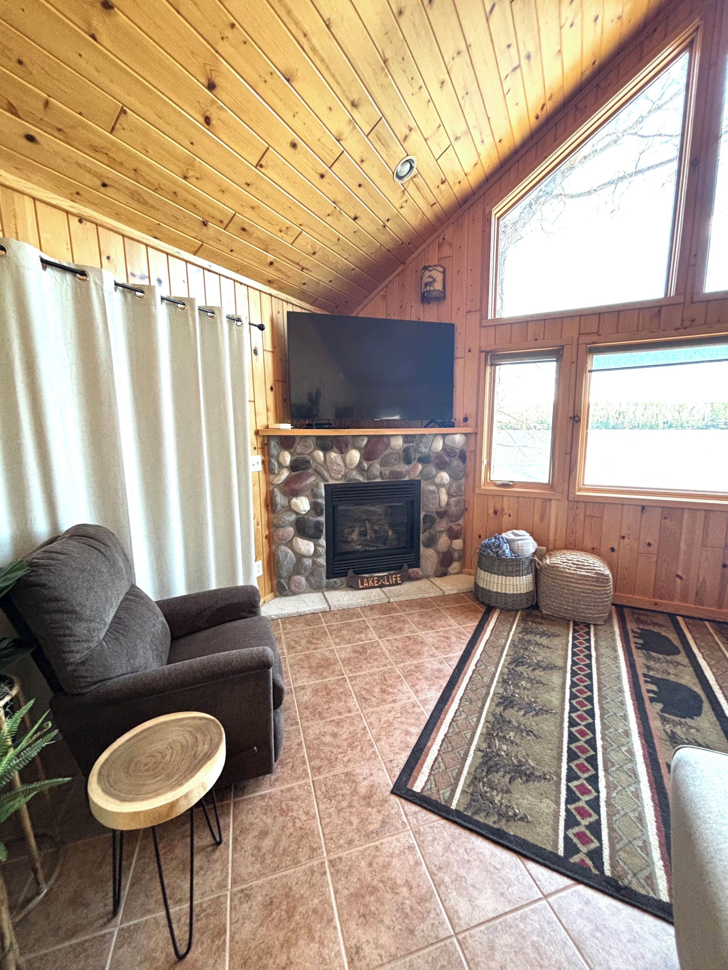 A cozy living room with a stone fireplace, a flat-screen TV mounted above it, and a large window providing natural light.