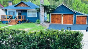A two-story house with a blue exterior, a front porch with a bench, and a garage with two wooden doors. - The Emerald Cottage/Main Street/Garage (Deadwood)