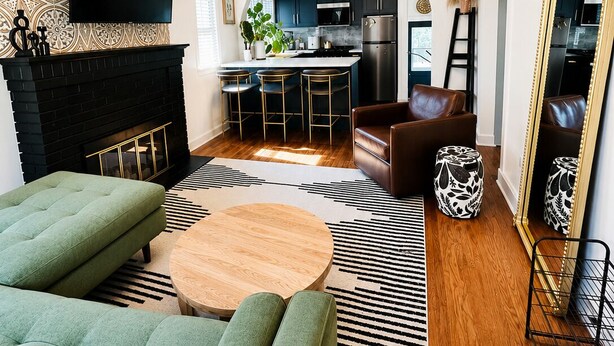 A modern living room with a fireplace, a sofa, a coffee table, and a kitchen in the background. - The Emerald Cottage/Main Street/Garage (Deadwood)