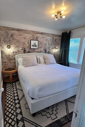 Un dormitorio con cama, mesita de noche, lámpara de pared y ventana con cortinas.