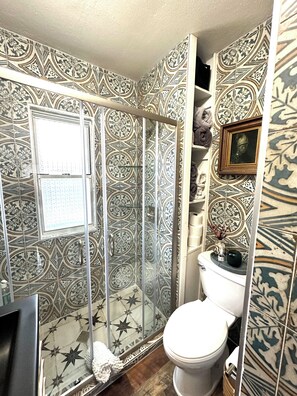 Un baño con baldosas estampadas, cabina de ducha de vidrio, inodoro y un cuadro en la pared.