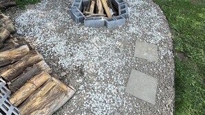 Un foyer circulaire entouré de gravier et de dalles de pierre, avec un banc en bois et une pile de bois de chauffage à proximité.
