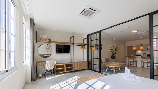 Une chambre d’hôtel moderne équipée d’un lit, d’un bureau, d’une chaise, d’une petite table, d’une plante en pot et d’une porte-fenêtre coulissante donnant sur un coin cuisine.