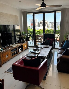 A modern living room with a flat-screen TV, a red sofa, a coffee table, and a balcony with a view of buildings. - Darwin City Apartment 
Large Garden Balcony with Uninterrupted Views... (Darwin City)