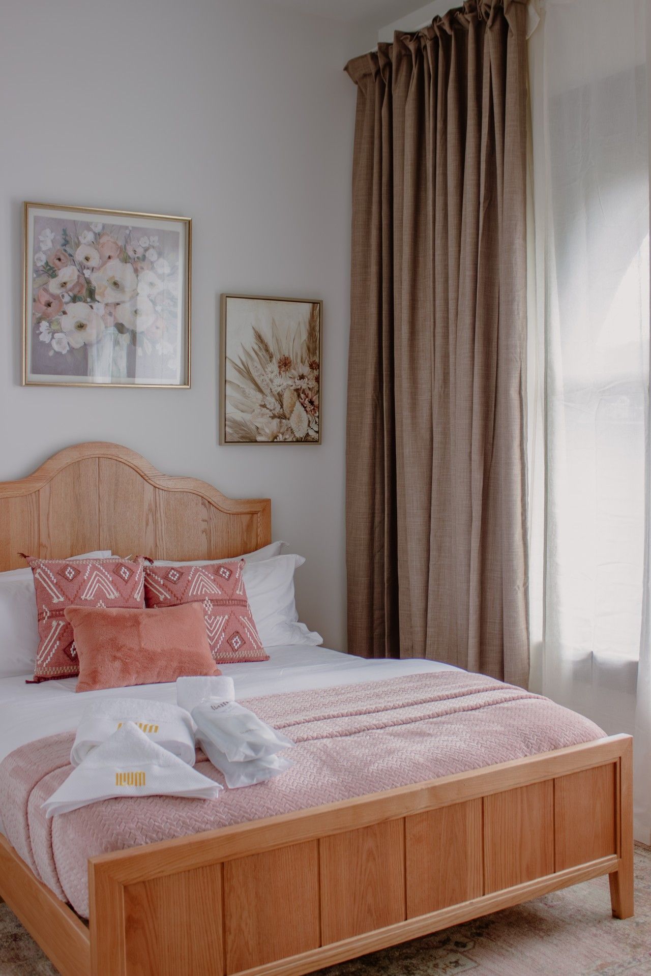 A bedroom with a wooden bed, two framed floral artworks, and a large window with curtains.