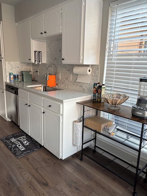 Een moderne keuken met witte kasten, een inbouwvaatwasser, een koffiezetapparaat en een raam met zonwering.