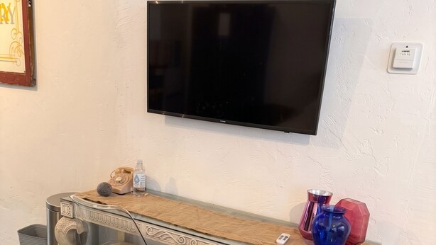 Een kamer met een wandtelevisie, een houten tafel met twee stoelen, een prullenbak en een kleed op de vloer.