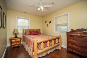 Un dormitorio con cama, mesita de noche, cómoda, ventilador de techo y dos ventanas con persianas.