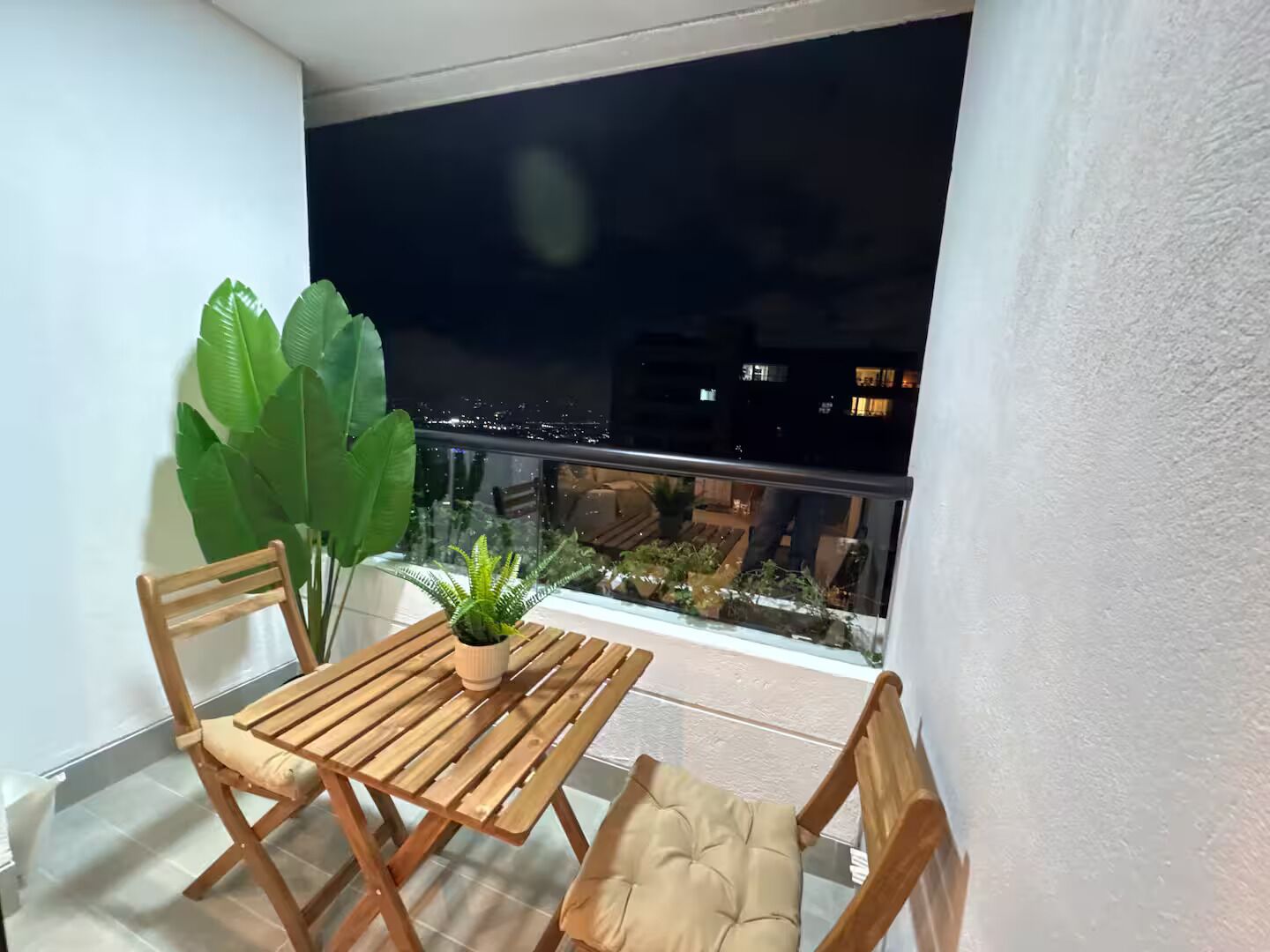 A balcony with a wooden table and chairs, a potted plant, and a view of a cityscape at night.