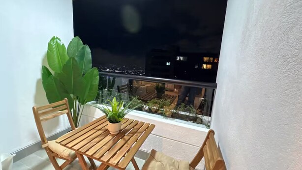 Un balcón con mesa y sillas de madera, una planta en maceta y vista a un paisaje urbano nocturno.