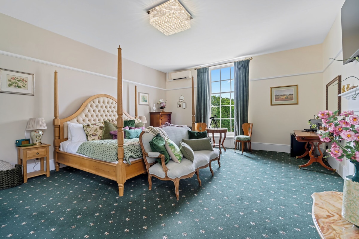 A bedroom with a four-poster bed, a sitting chair, a desk, and a large window with curtains.