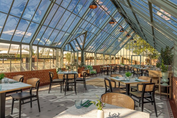 Un restaurante estilo invernadero con mesas, sillas, ventanas amplias y plantas en macetas.