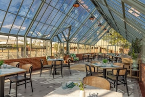 Um restaurante em estilo estufa, com mesas, cadeiras, janelas amplas e plantas em vasos.