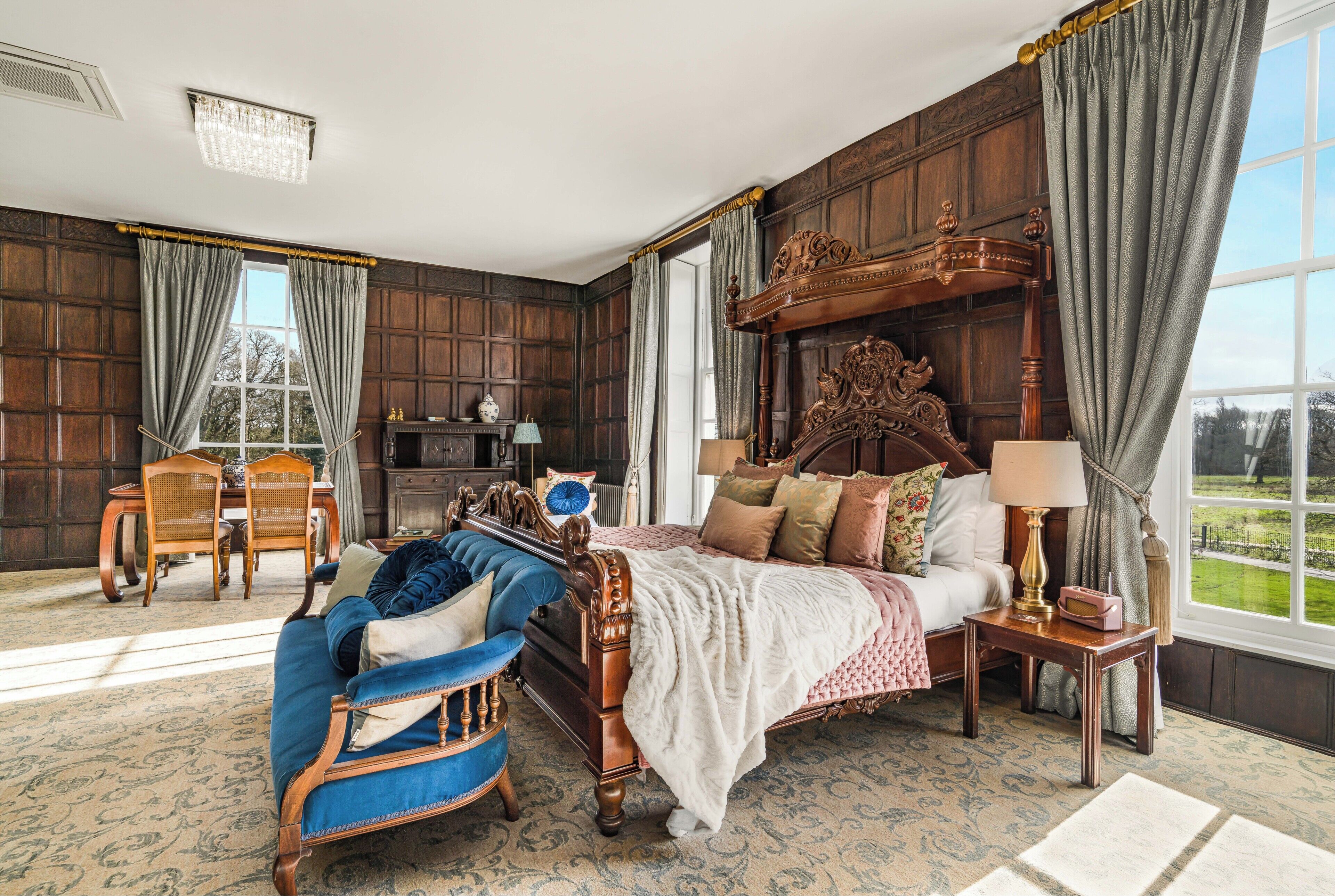 Une chambre avec un grand lit, un canapé bleu, une tête de lit en bois, une table de chevet avec une lampe et une vue sur l’extérieur.
