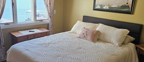 A bedroom with a bed, a ceiling fan, a window with curtains, a painting on the wall, and a wooden nightstand.
