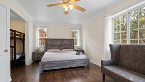A bedroom with a wooden floor, a bed with a grey headboard, a grey sofa, a ceiling fan, and two windows. - Paramount Escape Big Bear-Close to Everything -Tesla Universal EV- Foos Ball (Big Bear Lake)