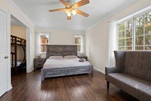 Ein Schlafzimmer mit Holzboden, einem Bett mit grauem Kopfteil, einem grauen Sofa, einem Deckenventilator und zwei Fenstern.