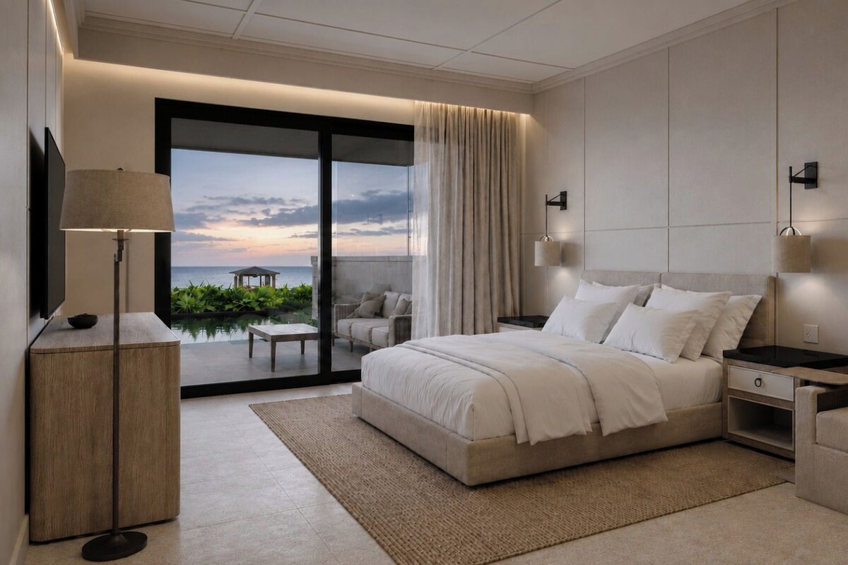 A modern bedroom with a large bed, a nightstand, a sofa, and a view of the ocean through a sliding glass door.