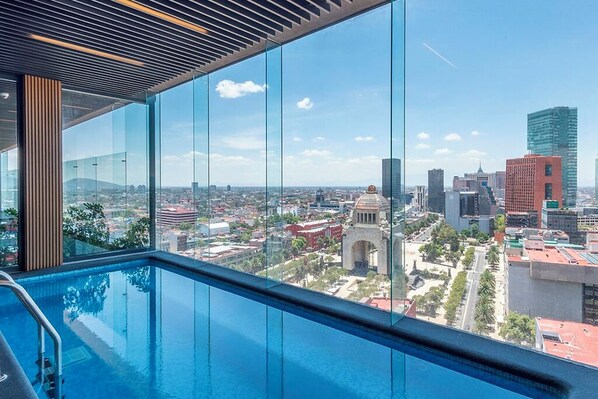 A modern hotel room with a large window overlooking a cityscape, a pool, and a view of a historic arch. - Central loft with luxury amenities, close to the historic center and El Ángel (Ciudad de México)