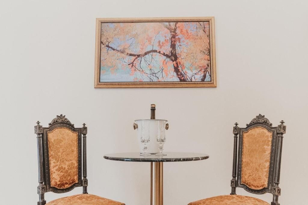 A room with two ornate chairs, a small round table with a clear glass top, and a framed painting of a tree on the wall.