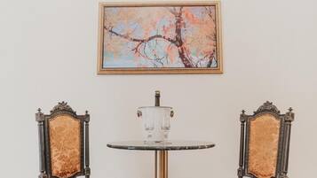 A room with two ornate chairs, a small round table with a clear glass top, and a framed painting of a tree on the wall.