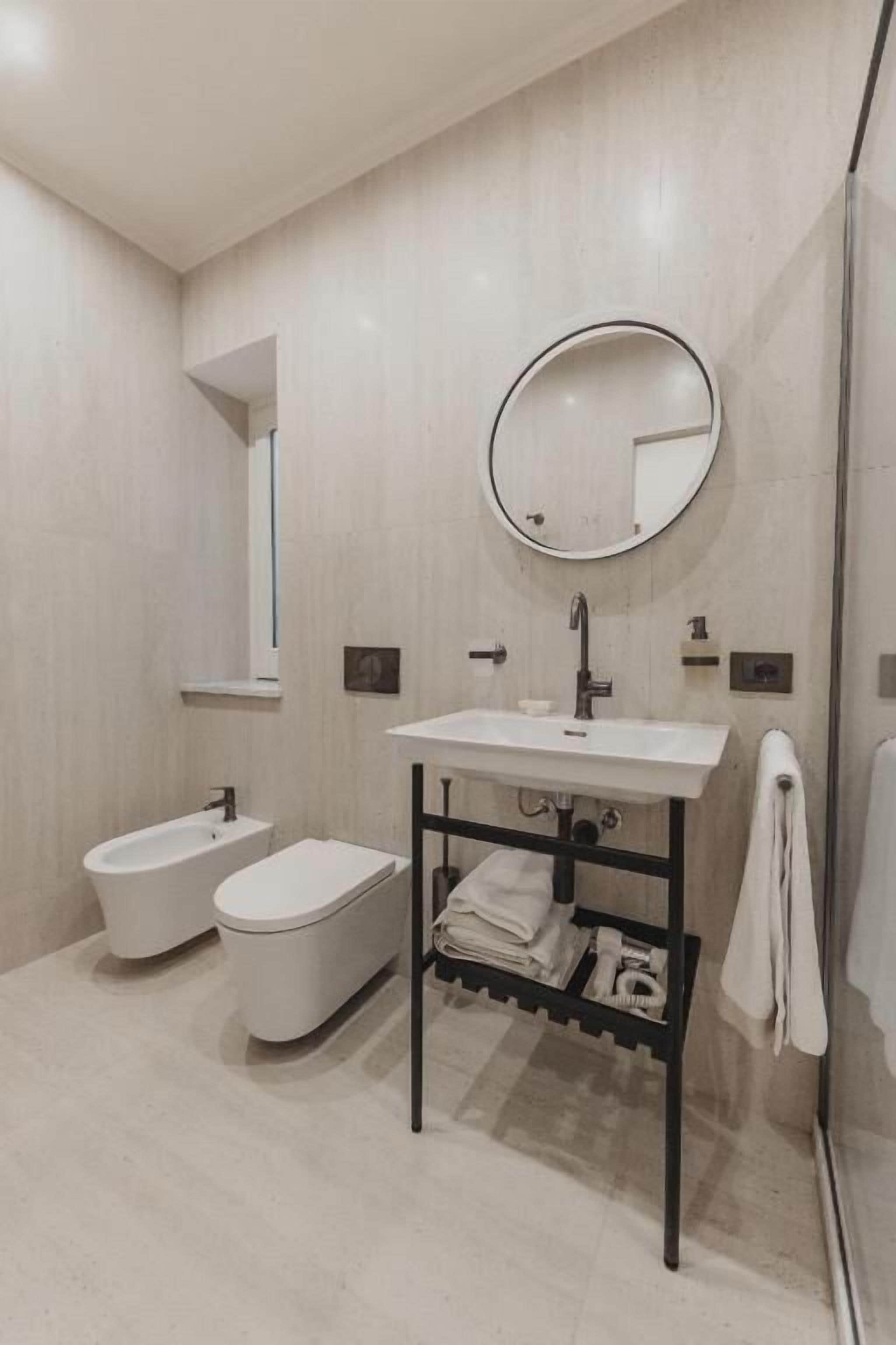 Un bagno moderno con un lavandino bianco, uno specchio rotondo, un bidet e un portasciugamani.