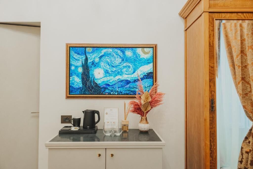 A room with a painting of a swirling night sky, a coffee maker, a vase with dried flowers, and a wooden cabinet.