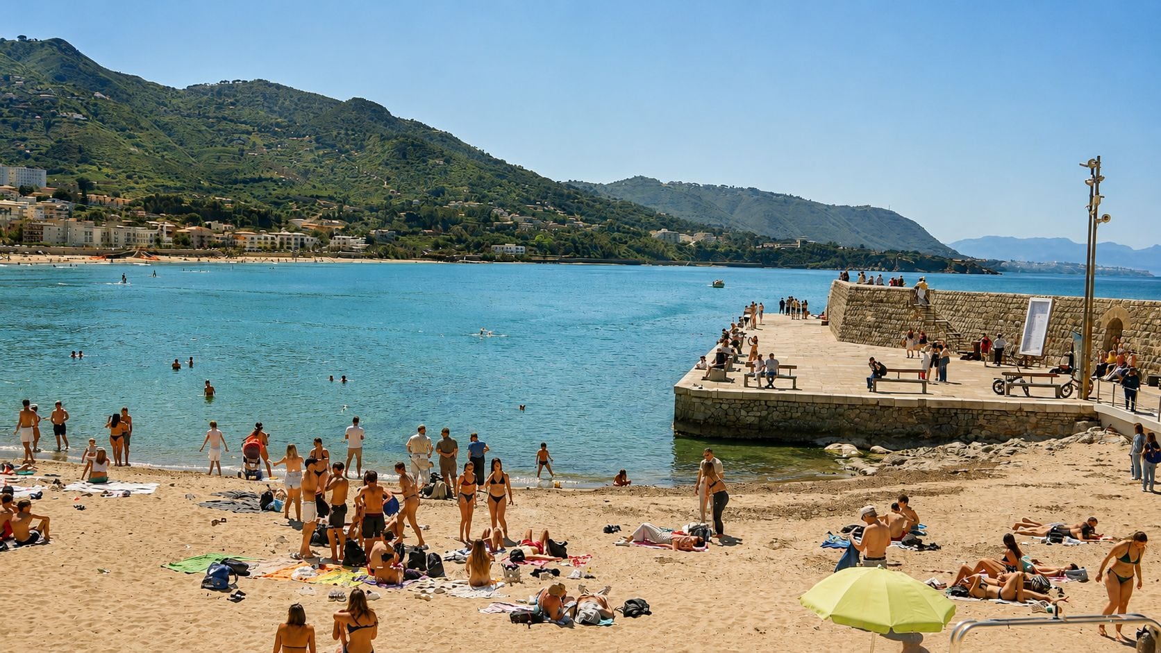 A beach with people sunbathing and swimming, a stone pier, and hills in the background.
