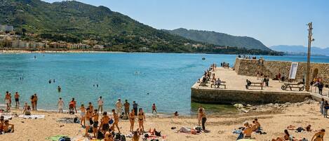 A beach with people sunbathing and swimming, a stone pier, and hills in the background.