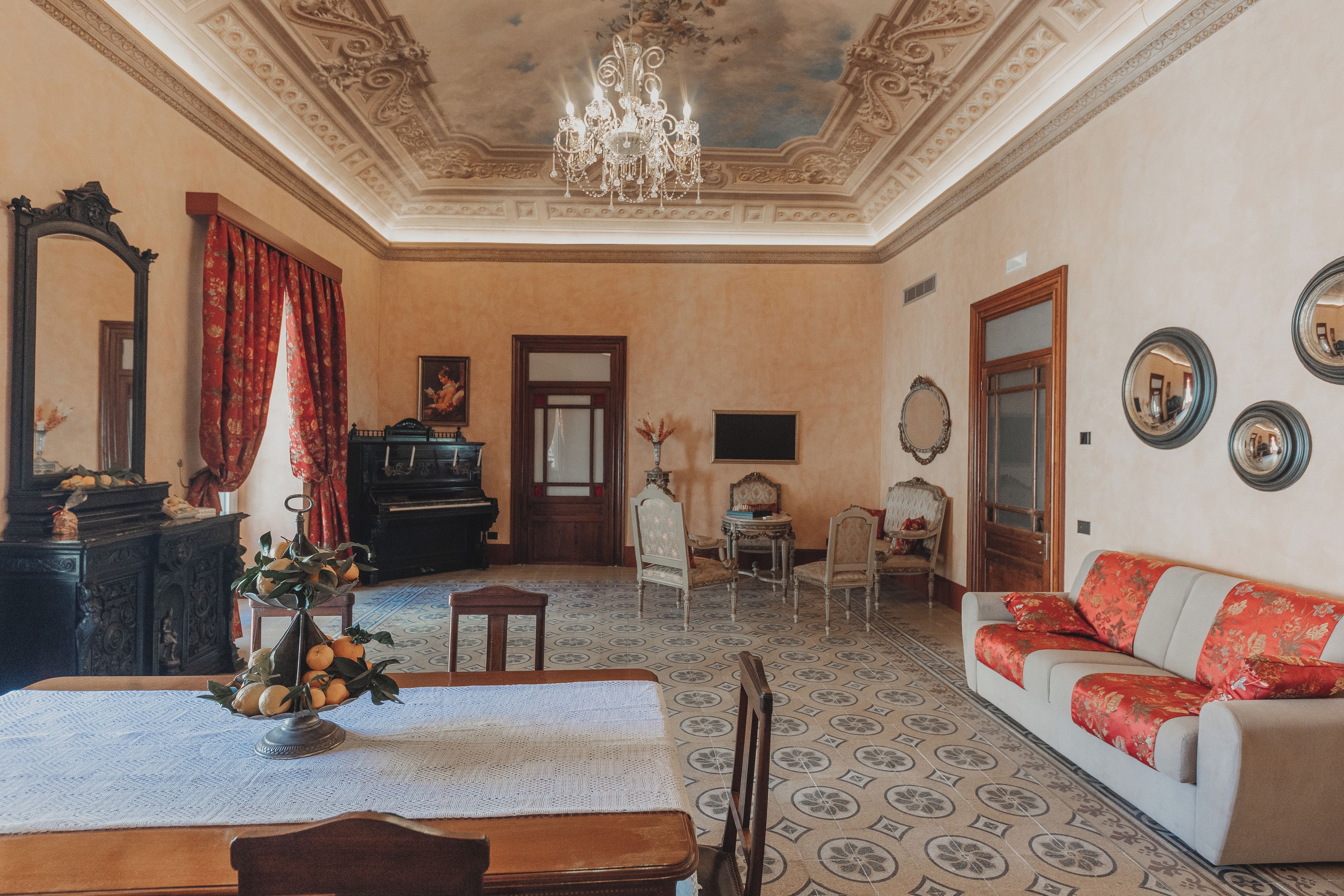 A room with a piano, a dining table with a vase of flowers, a sofa with red cushions, and a chandelier.