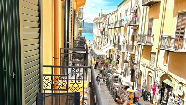 Una calle estrecha con balcones, mesas al aire libre y tiendas.