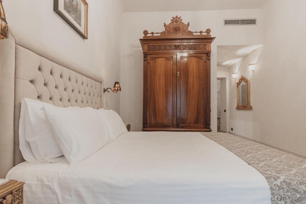 Un dormitorio con cabecera capitonada, un amplio armario de madera, cama con ropa blanca y alfombra estampada.