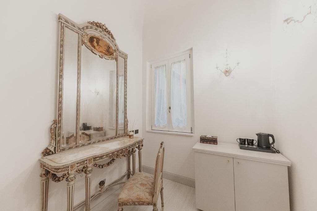 Una habitación con un espejo grande y decorativo, un tocador con una silla, un armario y una ventana con cortinas.