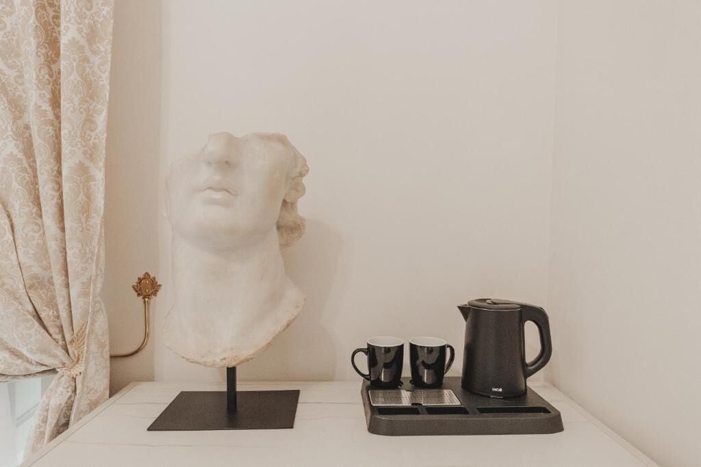 A bust sculpture on a stand, two black mugs, and a black coffee pot on a tray.