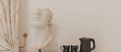 A bust sculpture on a stand, two black mugs, and a black coffee pot on a tray.