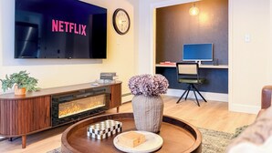 Una sala de estar con un televisor mostrando 'NETFLIX', una chimenea, una mesa de centro con objetos decorativos, un escritorio con una computadora y una silla.
