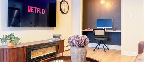 A living room with a TV displaying 'NETFLIX,' a fireplace, a coffee table with decorative items, a desk with a computer, and a chair.