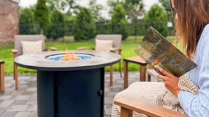 Una persona leyendo un libro en una mesa redonda al aire libre con una chimenea en el centro, rodeada de sillas y un muro de ladrillo al fondo.