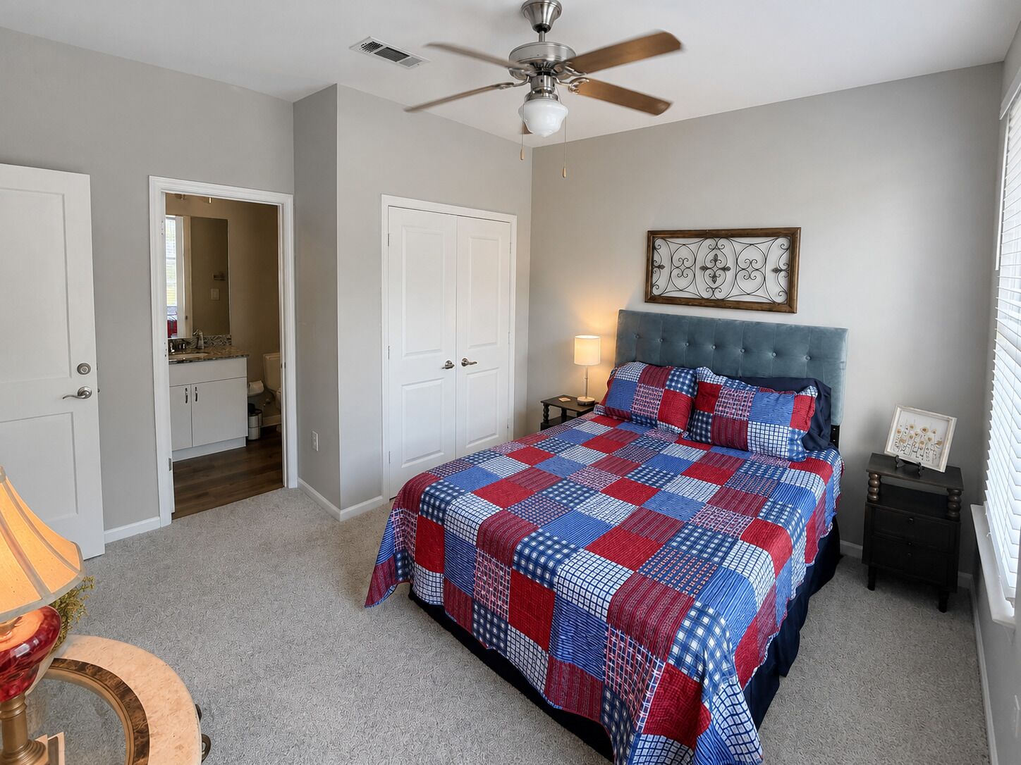 A bedroom with a bed, a ceiling fan, a nightstand, a lamp, a framed picture, and a door leading to a bathroom.