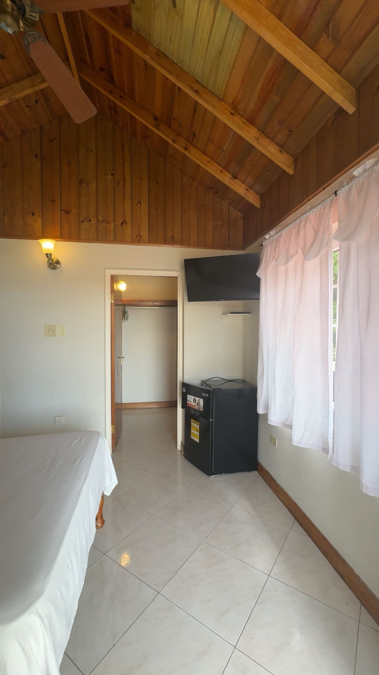 Une chambre avec un lit, un petit réfrigérateur noir, une télévision et une fenêtre avec des rideaux.
