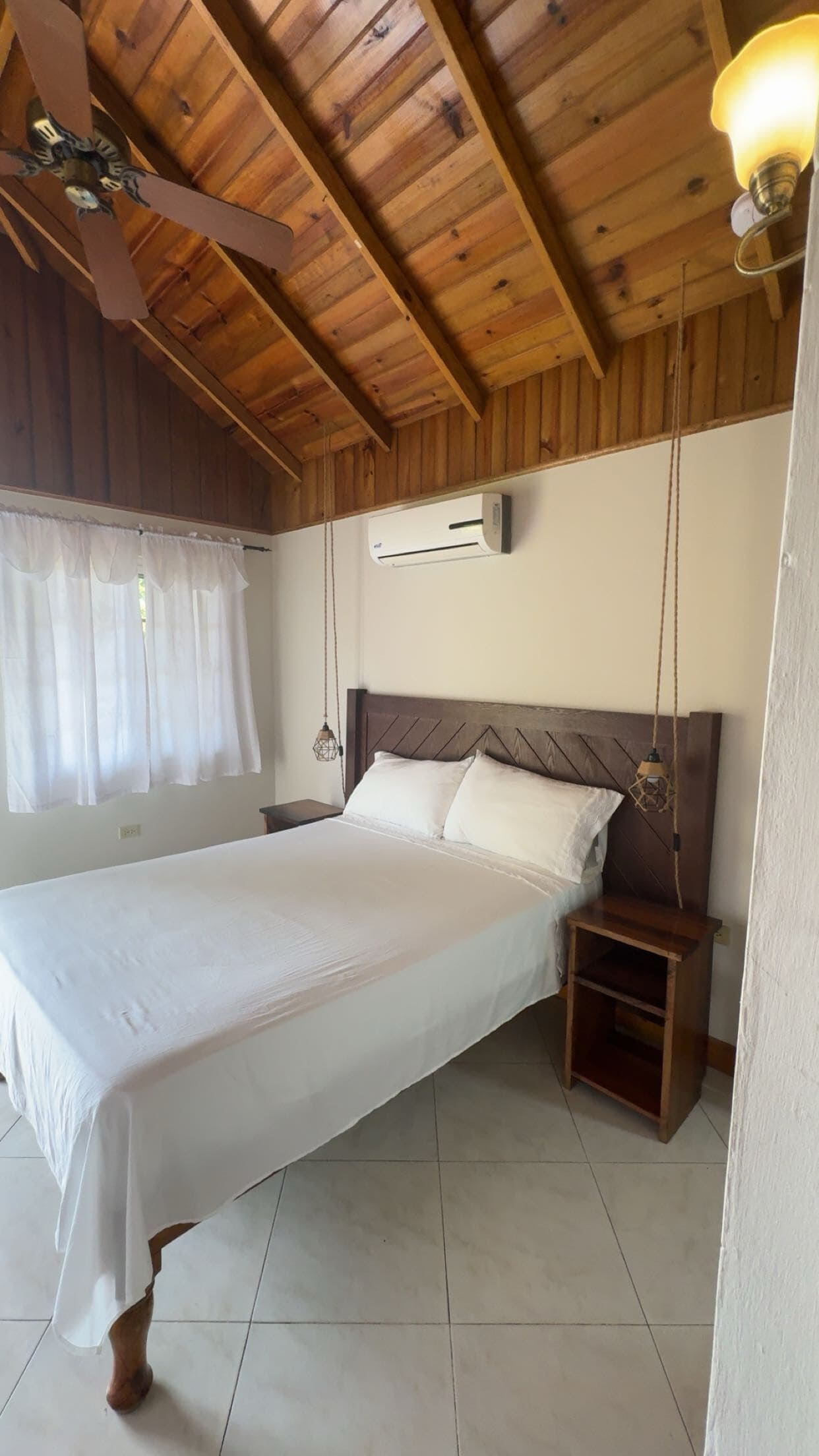 A bedroom with a wooden ceiling, a bed with white bedding, a nightstand, and a wall-mounted air conditioner.