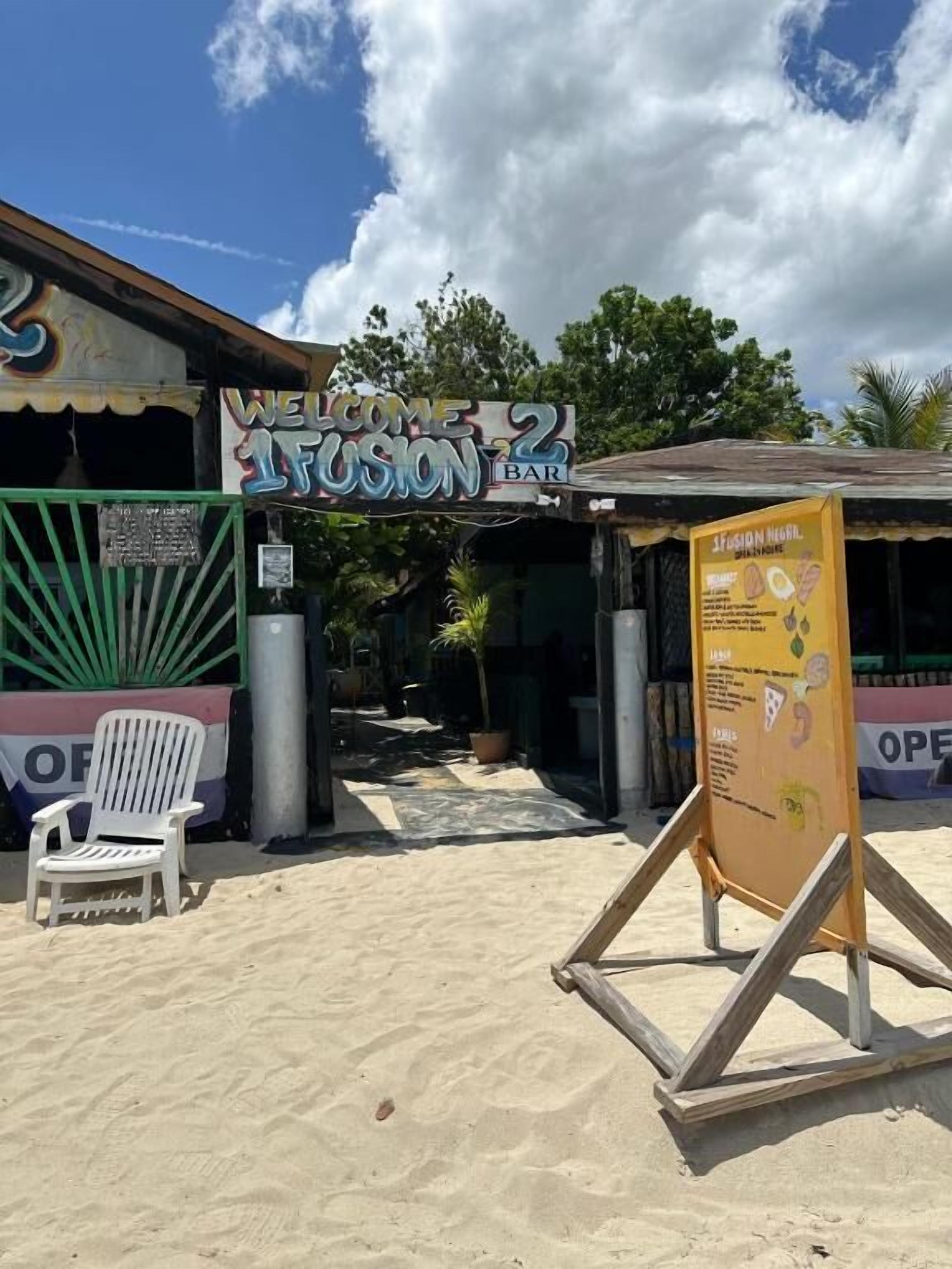 Um bar à beira-mar chamado 'Welcome to Fusion Bar', com uma placa, uma cadeira branca e uma estrutura de madeira.