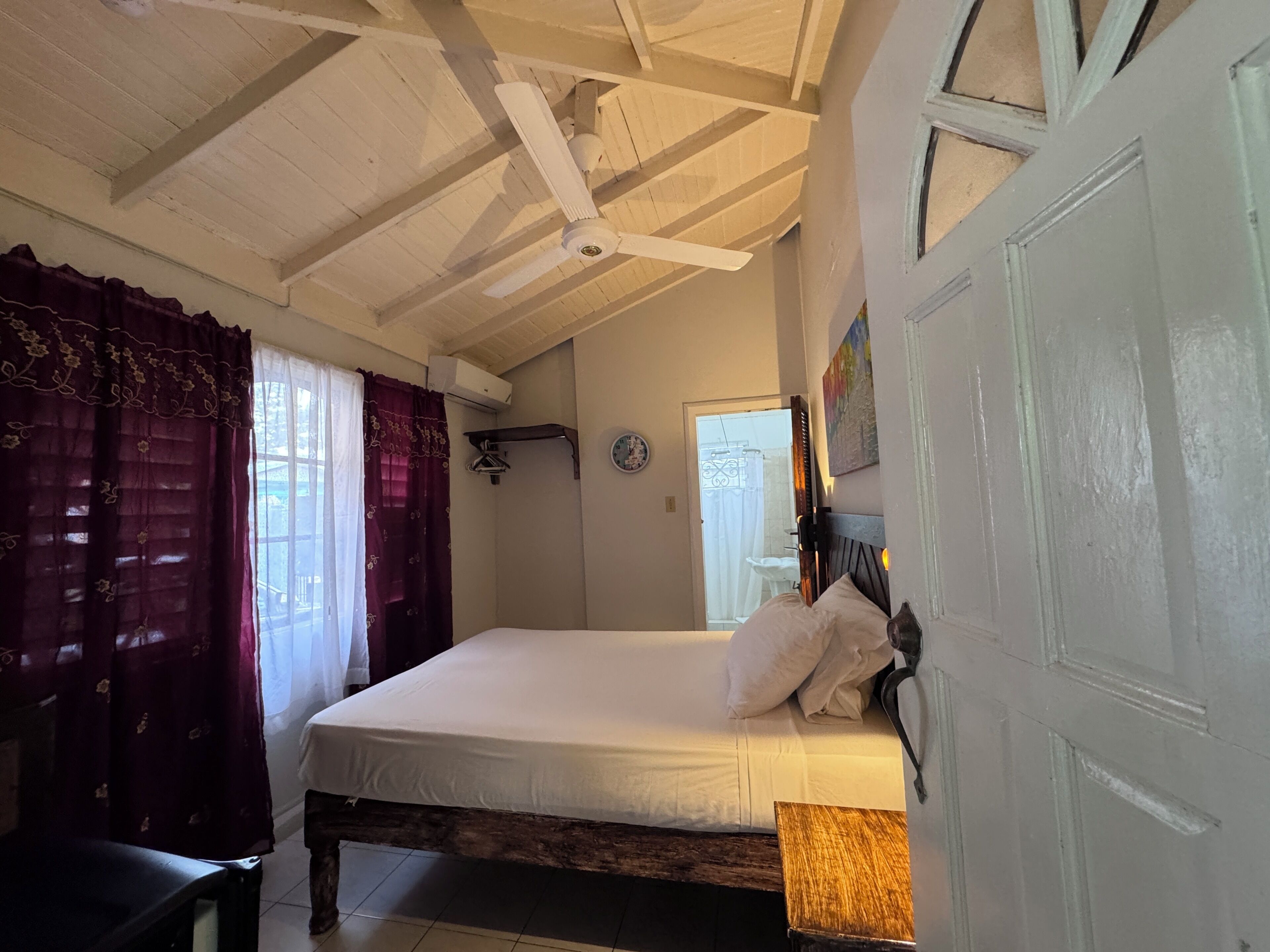 Un dormitorio con cama, techo de madera, puerta blanca y ventana con cortinas.