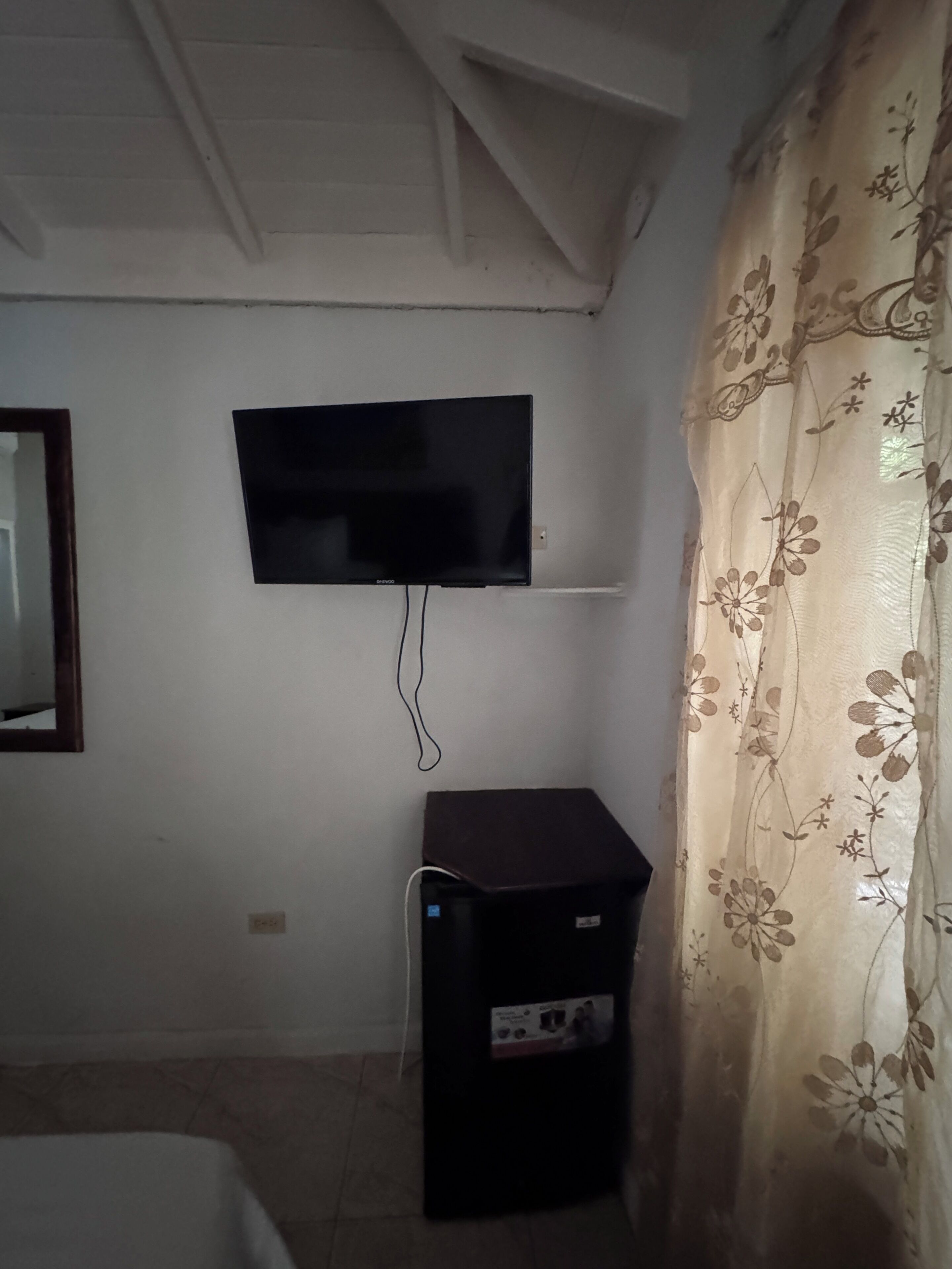 Een kamer met een aan de muur bevestigde televisie, een kleine zwarte speaker en een bloemenprint gordijn.