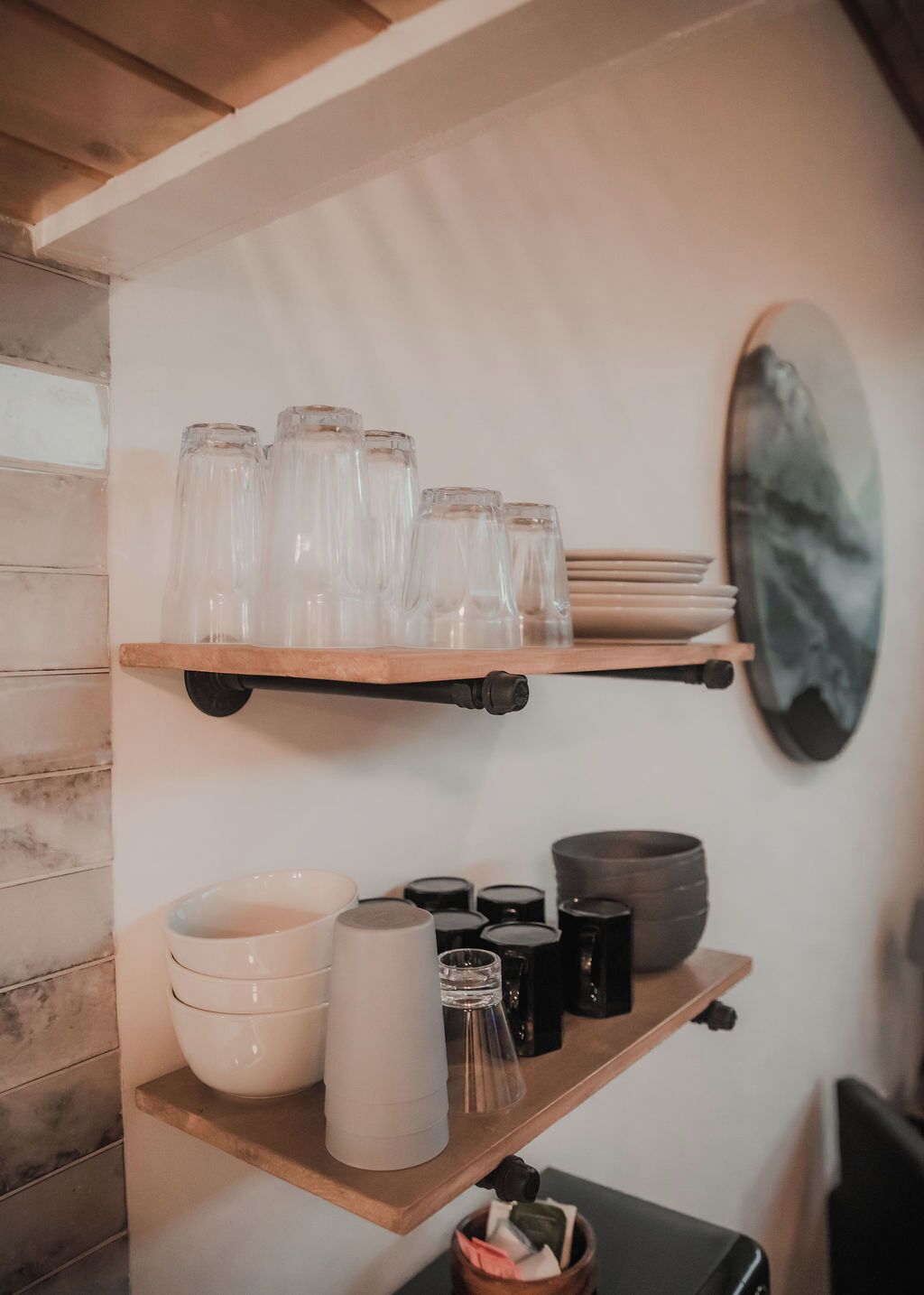 A kitchen with wooden shelves holding glassware, bowls, and black containers.
