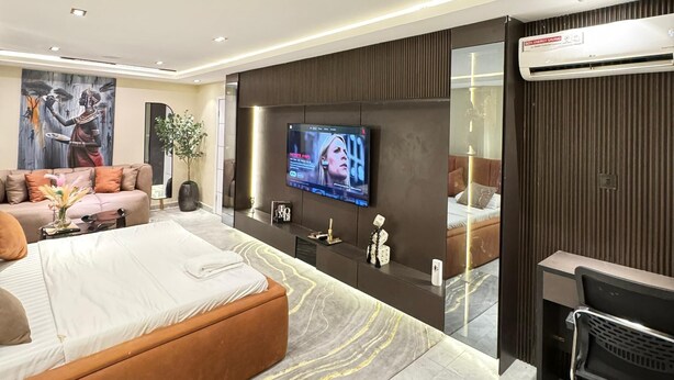 A modern hotel room with a large bed, a TV mounted on the wall, a sofa, and a desk area. - Classic 1 bedroom apartment  (Lekki)