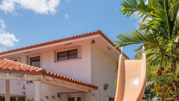 A pool with a slide, a house with a tiled roof, and palm trees. - La  Casona (Miami)