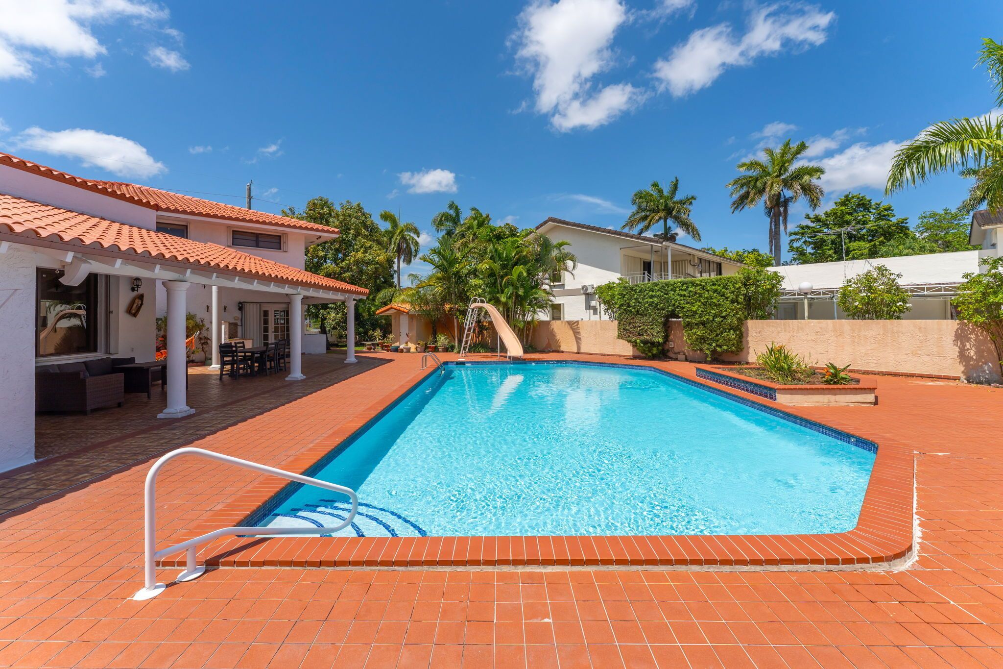 A swimming pool with a tiled deck, a covered seating area, and palm trees in the background.