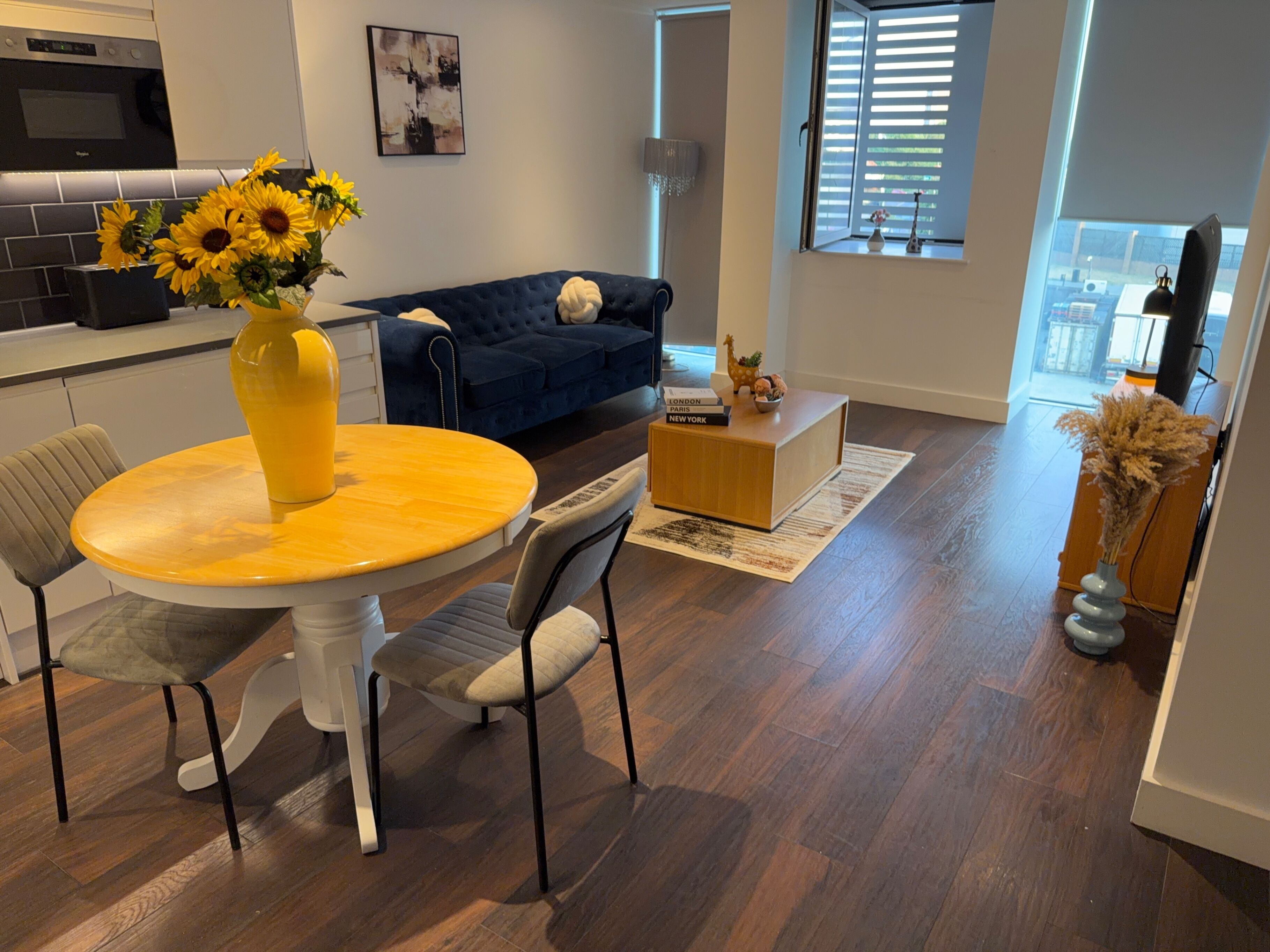 A modern living room with a dining table, chairs, a blue sofa, a coffee table, and a vase with sunflowers.